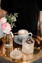 Woman Holding Bronze Serving Tray With Client's Order: Sweet Macaroon, Cup Of Coffee And Small Flowers Bouquet In Transparent Vase Nearby