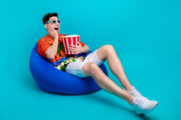 Canvas Print - Full size photo of ecstatic man dressed print shirt hold popcorn in lay on pouf staring empty space isolated on blue color background