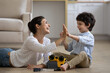 © fizkes - Excited Indian mother with 5s son repairing toy car, giving high five, sitting on warm wooden floor at home together, smiling laughing young mom and adorable boy child using screwdrivers, having fun