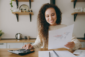 Young happy housewife woman of African American ethnicity wears casual clothes sweater use calculator studying read papers sit at table in light kitchen at home alone. Lifestyle cooking food concept.