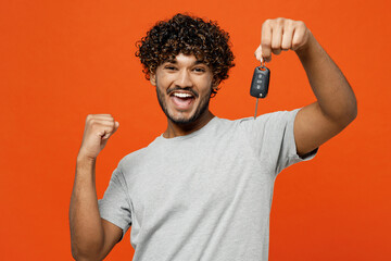 Young smiling happy fun Indian man he wears t-shirt casual clothes hold in hand car key fob keyless system do winner gesture isolated on orange red color background studio portrait. Lifestyle concept.