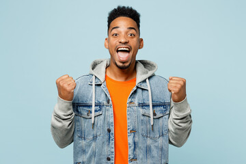 Wall Mural - Young man of African American ethnicity wear denim jacket orange t-shirt doing winner gesture celebrate clenching fists say yes isolated on plain pastel light blue background studio Lifestyle concept