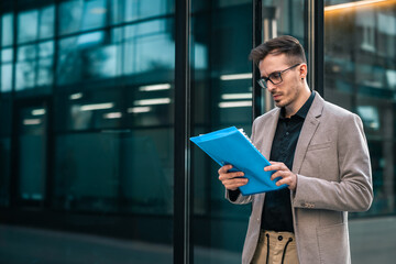 Serious businessman looking at paper documents in front of office building. Focused male manager checking reports before work while standing outdoors.