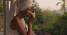 Starting new day. Close-up portrait of a happy woman with a cup of coffee or tea in a white towel after shower, standing on an open resort balcony against a nature background during a perfect morning