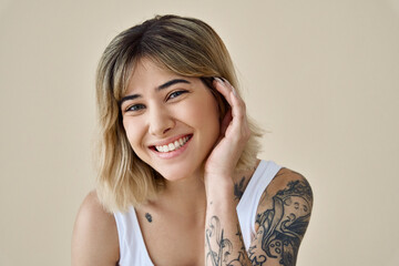 Wall Mural - Smiling blond pretty happy girl beauty female model with short blonde hair and tattoos looking at camera touching face pampering advertising skin care isolated at beige background. Portrait.