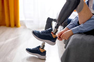 Wall Mural - rehabilitation after leg amputation, man getting used to prosthesis on legs, wearing sneakers sitting on bed in bright bedroom at home. difficulties in disabled people life, physical disability