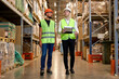 © alfa27 - two engineers in safety helmets and green uniforms walking among shelves with goods in warehouse talking, checking goods. logistic and business export, distribution and inventory concept