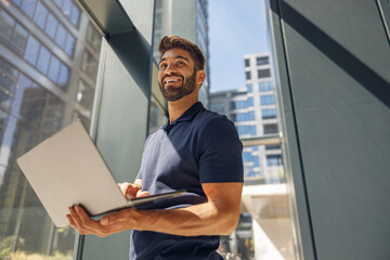 Handsome smiling freelancer working on laptop standing in coworking near window and looks away