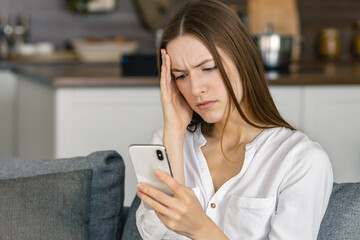 Sad woman holding a mobile phone, received notification message with bad news sitting on couch in living room