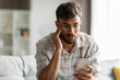 © Home-stock - Shocked indian guy looking at cellphone screen, sitting on couch at home, holding phone and touching his head, reading bad news or message, copy space