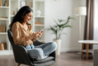 © Home-stock - Happy indian woman using cellphone, sitting in armchair, browsing internet or texting with friends, enjoying mobile communication at home, free space