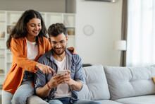Happy young indian couple using smartphone, wife pointing at cellphone screen, sitting on sofa in living room interior at home. New app ad, banner