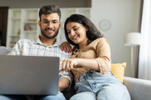Happy Indian Couple Using Laptop, Surfing Web, Woman Pointing At Screen, Reading Exciting News, Sitting On Sofa. Remote Entertainment, Online Life Concept