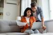 © Home-stock - Serious indian couple paying bills online, sitting on couch, using laptop, concentrated husband and wife checking papers and banking on Internet, copy space