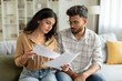 © Home-stock - Indian couple checking bills, reading documents, unhappy man and woman holding papers, counting monthly spendings, sitting on sofa at home