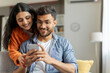 © Home-stock - Cheerful indian couple using cellphone, shopping online or sharing social media, spouses sitting on sofa at home interior, woman embracing her husband looking at cellphone screen