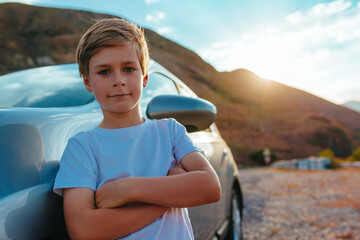 Wall Mural - Cut boy stands by the car in the mountains at sunset, travel concept