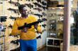 African-american man standing in salesroom of gun shop and selecting assault rifle.