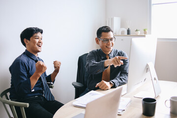 Smiling mature Asian businessman pointing at computer with partner clenched fist showing victory hands gesture. Business people celebrating target achievement