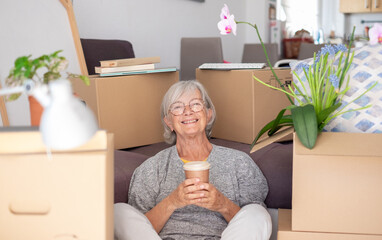 Happy senior woman involved in moving house sitting among cardboard boxes takes a break with a coffee, concept of moving, retirement, new life, buying, renting, apartment, house