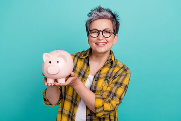 Photo of charming grandmother hold pig bank collect money dressed stylish checkered yellow outfit isolated on aquamarine color background