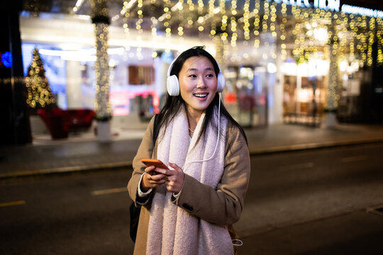 Young relaxed lady texting and wearing headphones with christmas lights behind. Happy girl listening to music at the street during winter using her phone.