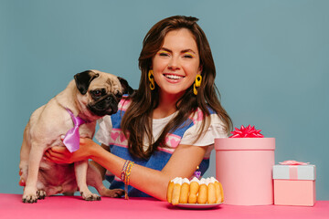 Wall Mural - Joyful woman petting cute dog and celebrating birthday while sitting at the pink desk on blue background