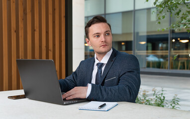 Young businessman in a suit and tie working on a laptop in a street cafe near the business center. Man working on laptop with blank back panel without stickers