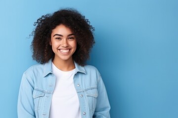 Confident female high school student wearing oversized denim jacket and white t - shirt, standing against blue background.