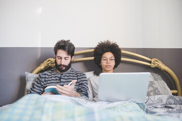 Young multiethnic couple bed using pc reading book