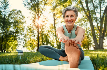 Wall Mural - Lady performing stretching exercise. Portrait of cheerful lady with trendy hairstyle training outdoors on yoga rug and reaching for toe. Positive senior woman looking at camera and smiling.