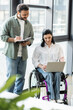 © LIGHTFIELD STUDIOS - happy indian man taking notes near disabled businesswoman on wheelchair using laptop in office