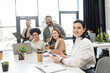 © LIGHTFIELD STUDIOS - happy businesswoman holding tablet and looking at camera near multicultural colleagues on background