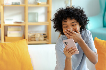 Wall Mural - Excited happy young black african american woman holding cell phone laughing feeling joy getting mobile message. Overjoyed girl laughing aloud sitting on couch watching funny video reading news