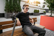 © phoenix021 - Relaxed young modern man sitting on bench in front of modern residential building , headphones on