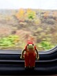 A toy frog traveler with a red backpack looks near the window in the train.  There are autumn trees outside the window.