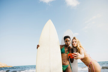 Wall Mural - Female surfers with their boards taking a selfie on the beach. Two women in bikinis having fun on a surfing holiday.