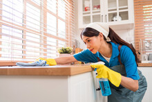 Professional Asian woman cleaning service wearing yellow rubber gloves, using a rag to wipe with spraying liquid detergent on the wooden top kitchen counter at home. Housekeeping cleanup, cleaner.