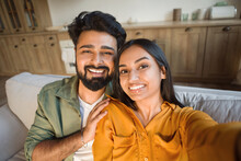 Closeup Portrait Of Cheerful Indian Couple Taking Selfie, Resting On Sofa At Home And Smiling, Posing In Living Room