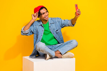 Wall Mural - Full size photo of nice young guy sitting white podium take selfie gadget wear trendy jeans outfit isolated on yellow color background