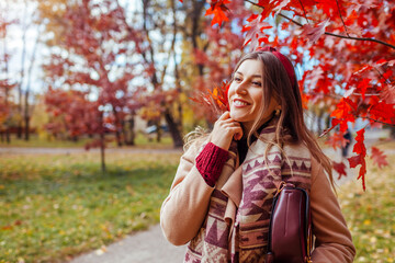 Wall Mural - Happy young woman walking in autumn park among red trees. Female fall fashion. Stylish girl wearing coat holding purse