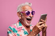 © arhendrix - Stylish cheerful elderly man in glasses rejoices and looks at smartphone screen on pastel