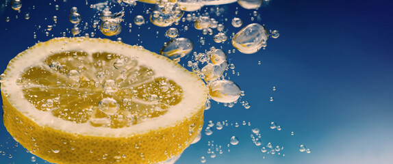 Wall Mural - Underwater of freshly squeezed sweetened lemonade which raw lemons slice falling in soda water against dark blue or black background. Close up lemonade or Highball lemon cocktail cold refreshing drink