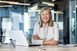 © Liubomir - Portrait of senior mature gray-haired businesswoman inside office, female boss sitting at desk and looking at camera with crossed arms, experienced financier with laptop at workplace.