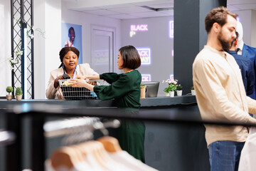 Wall Mural - Black Friday and retail industry. Young Asian woman customer standing at clothing store checkout counter giving clothes to friendly cashier, buying garment at discounted prices during seasonal sales