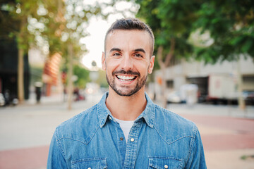 Close up portrait of handsome guy with perfect white teeth smiling and looking at camera standing outdoors. Front view of young man with friendly and positive expression. Happy adult male staring