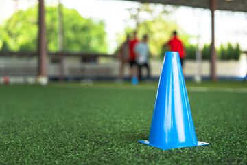 Wall Mural - A blue obstracle cone which is placed on football turf ground, using for the movement and speed training session, photo with group of player as blur background. Sport practice activity scene.