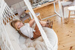 Little girl rides on swing on macrame chair at home. Rustic style in the interior. Toddler is wearing mustard corduroy pants and boots. Child is playing and fooling around. Cozy atmosphere. Copy space