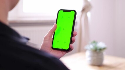 Slow motion of unrecognizable young man sitting on the couch in a living room and using his smartphone. Scrolling on a green screen with tracking markers, browsing on the internet. App template. 4K