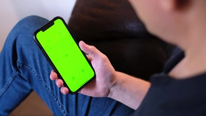 Unrecognizable young man sitting on the couch in a living room and using his smartphone. Scrolling, swiping, touching on a green screen with tracking markers. Browsing on the internet. App template.